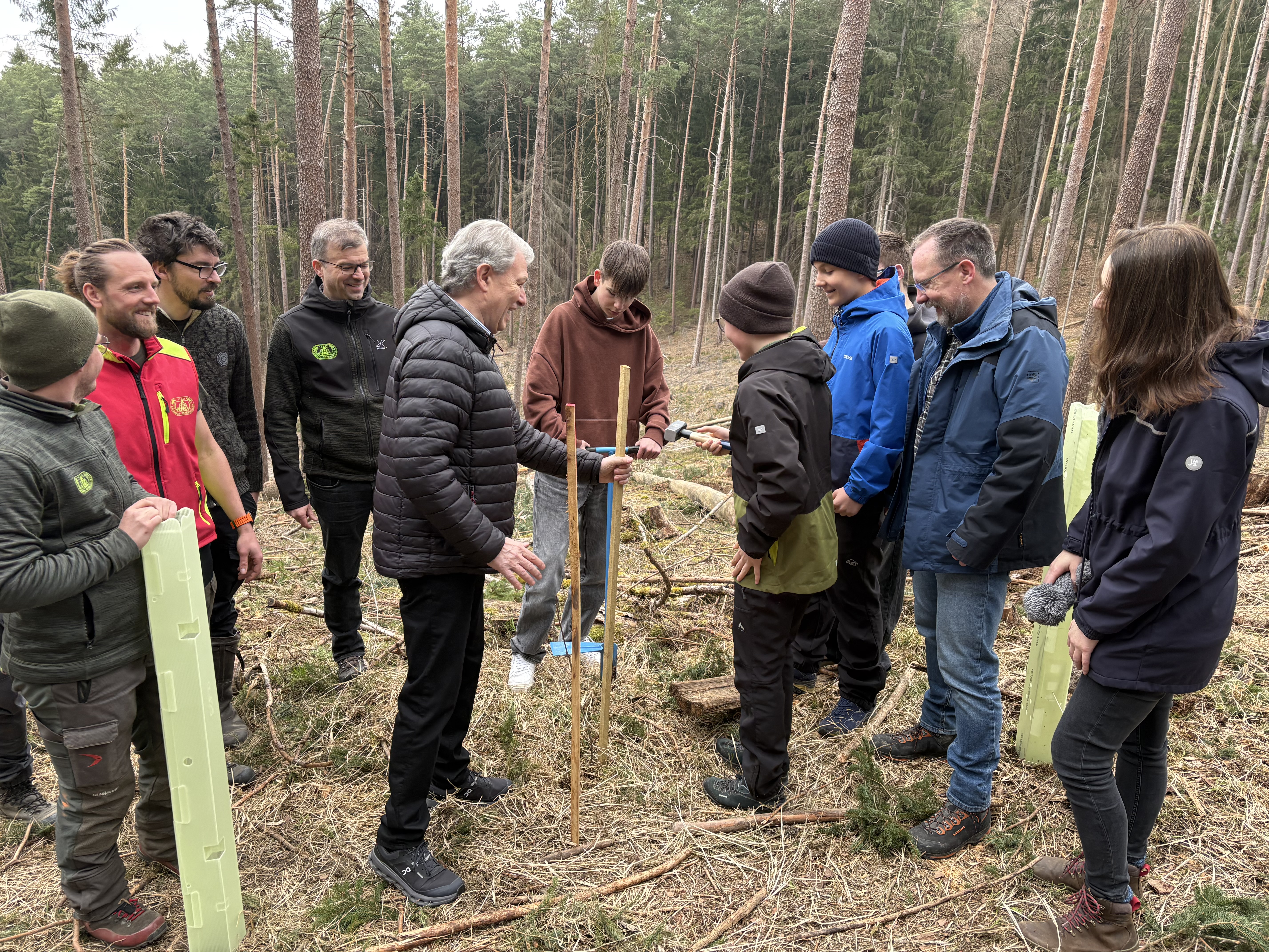 Das Bild zeigt einen Schüler mit Landrat Klaus Löffler beim Einschlagen einer Holzleiste, die als Halterung für den Pflanzenschutz dient. Außerdem sind noch Menschen mit auf dem Foto.