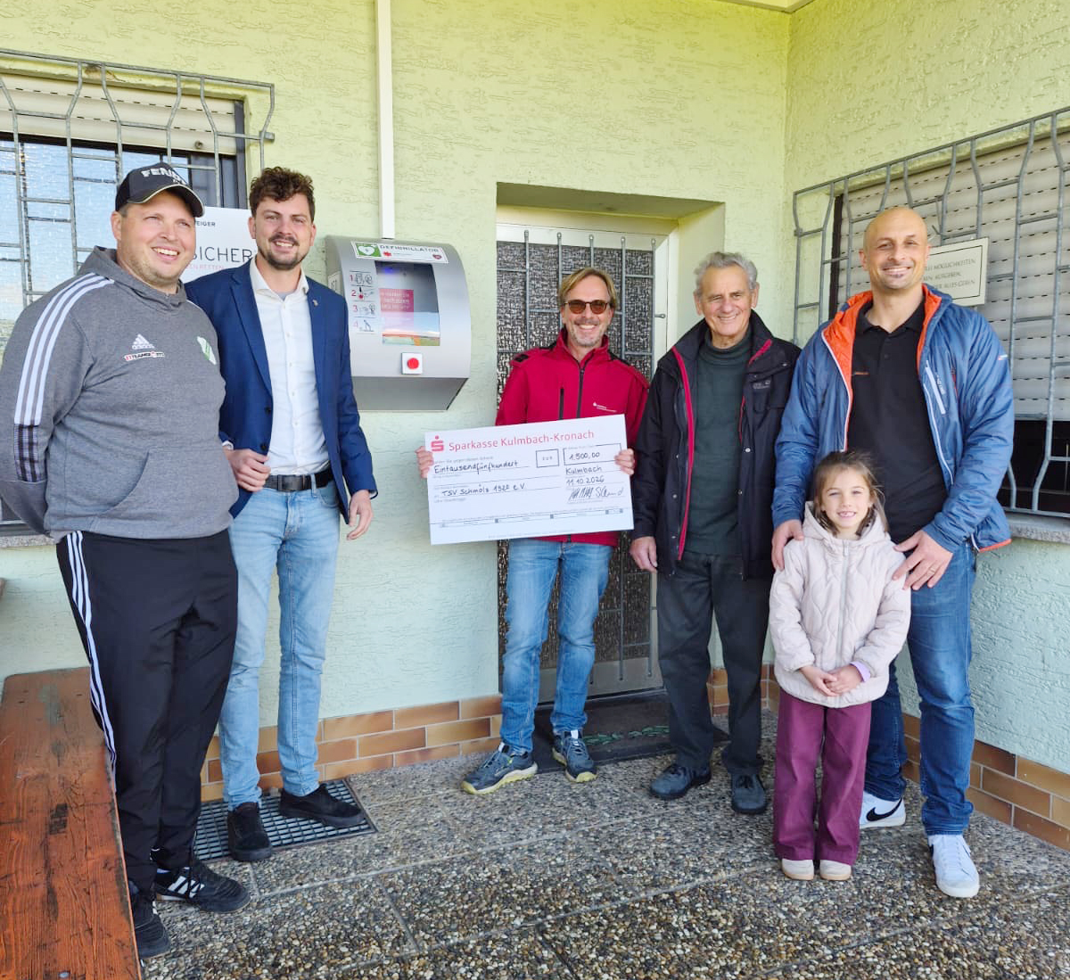 hier stehen Menschen vor einer Eingangstür und im Hintergrund ist der Defibrilator zu stehen